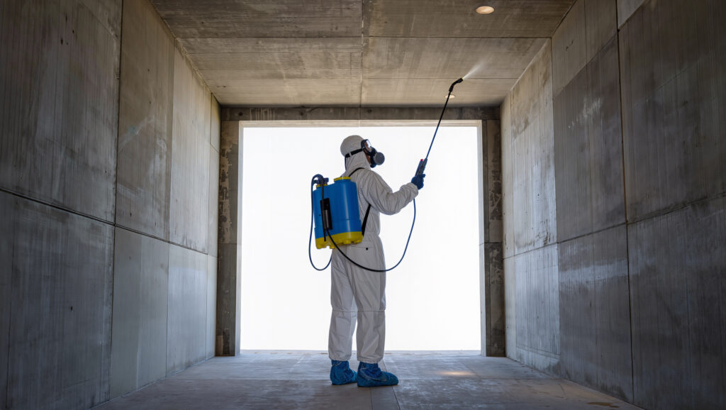 מדביר מקצועי מחברת ביו ישראל (Bio Israel) בציוד מיגון מלא וערדליים כחולים מבצע ריסוס תקרה בתוך מבנה בטון או חניון